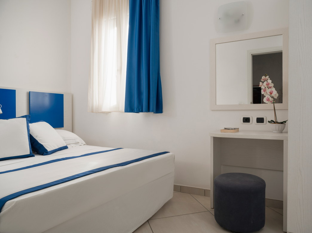Camera moderna con letto bianco e blu, specchio e finestra con tende blu.